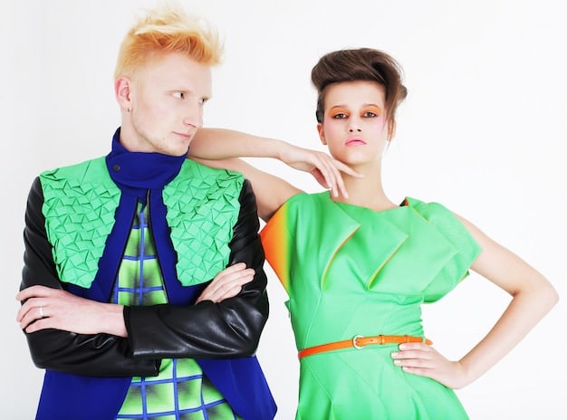 Two models, one male and one female, wearing identical oversized blazers in a bold color. The styling is androgynous, with minimal makeup and simple accessories. The background is a neutral studio setting, emphasizing the clothing.