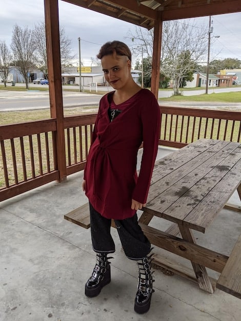 A woman with an apple-shaped body wearing an empire-waist dress with a V-neck, paired with dark leggings and ankle boots. The background is a trendy urban cafe.