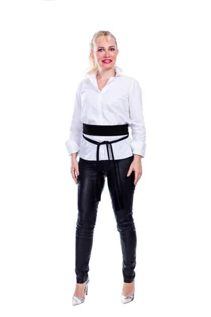 A woman wearing black tailored trousers, a white button-down shirt tied at the waist, and a denim jacket. She's accessorized with a neutral-colored scarf tied loosely around her neck and white sneakers.