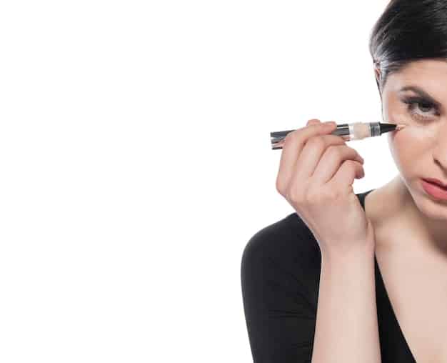 An overhead shot of a makeup artist applying black eyeliner with a fine brush to a model’s upper eyelid, closely following the lash line to enhance her smoky eye look.