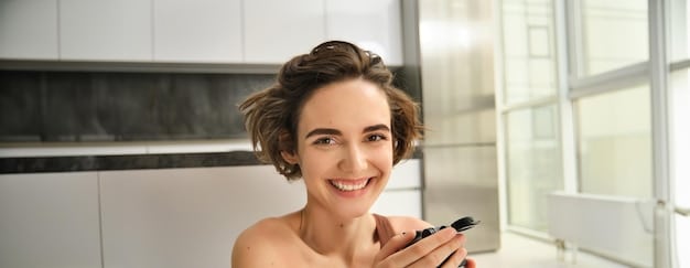 A woman applying cream blush to her cheeks with her fingertips. She is smiling and looking at the camera. The background is a well-lit bathroom.