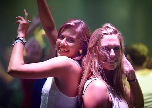 A medium shot of a group of friends at a music festival, all sporting unique and colorful makeup looks with glitter accents. The friends are smiling and posing for the camera, with the festival stage and crowd visible in the background.