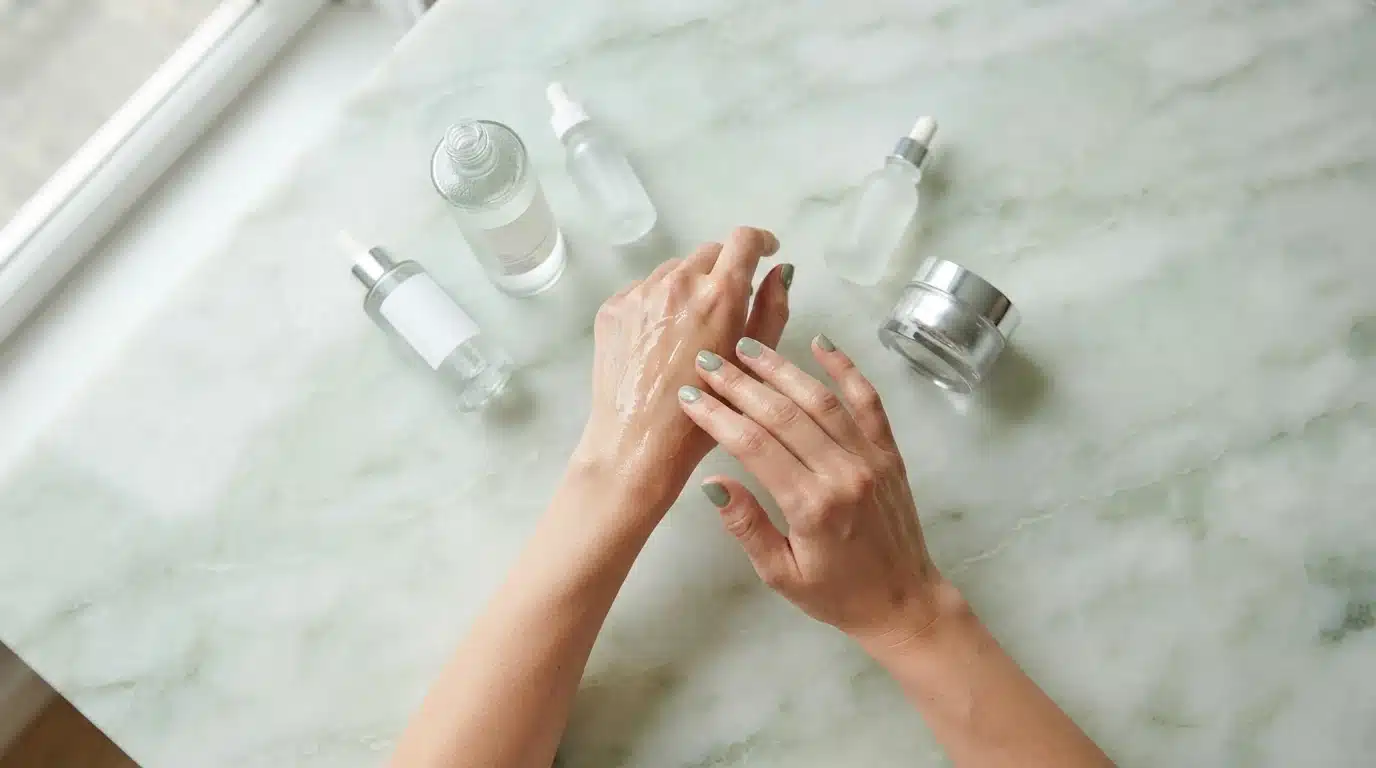 Overhead shot of hands applying hydrating serum, surrounded by skincare products on a green marble surface, demonstrating skin prep.